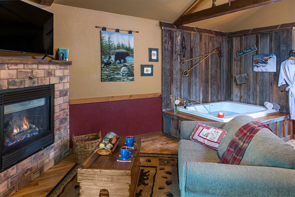 Cozy cabin interior featuring a brick fireplace with a roaring fire, a comfortable armchair, and an indoor Jacuzzi tub nestled in a corner with rustic wood paneling.