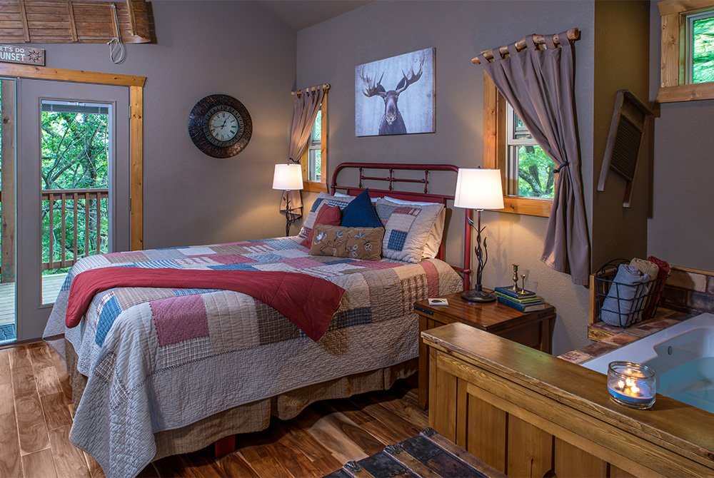 A cozy cabin bedroom featuring a red metal bed frame with a colorful patchwork quilt, a picture of a moose hanging on the wall, and an indoor Jacuzzi tub in the foreground.