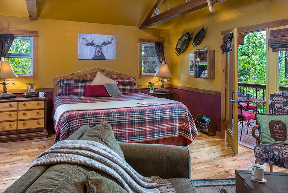 Cabin bedroom featuring a king-size bed with a plaid quilt, a large moose portrait on the wall, and glass doors opening onto a private deck with forest views.