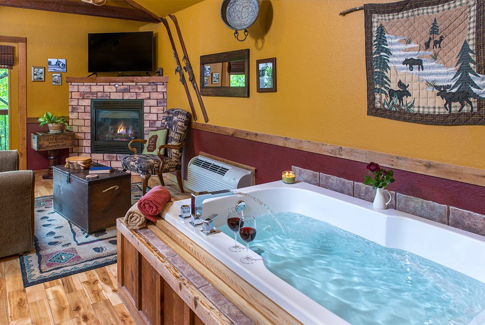 A spacious, bubbling Jacuzzi tub for two with wood-paneled siding, set in a cozy room featuring a brick fireplace and rustic mountain-themed wall decor.