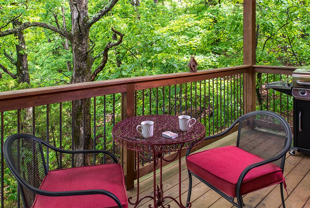 An outdoor wooden deck with two black chairs featuring red cushions, a small round metal table with two mugs, and a gas grill, overlooking a dense green forest.