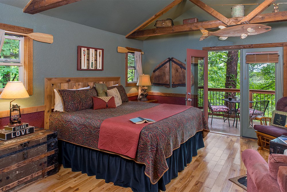 Bedroom interior with a king-size bed, featuring rustic fishing-themed decor like mounted oars and a large fish sculpture, with glass doors leading to a wooded balcony.