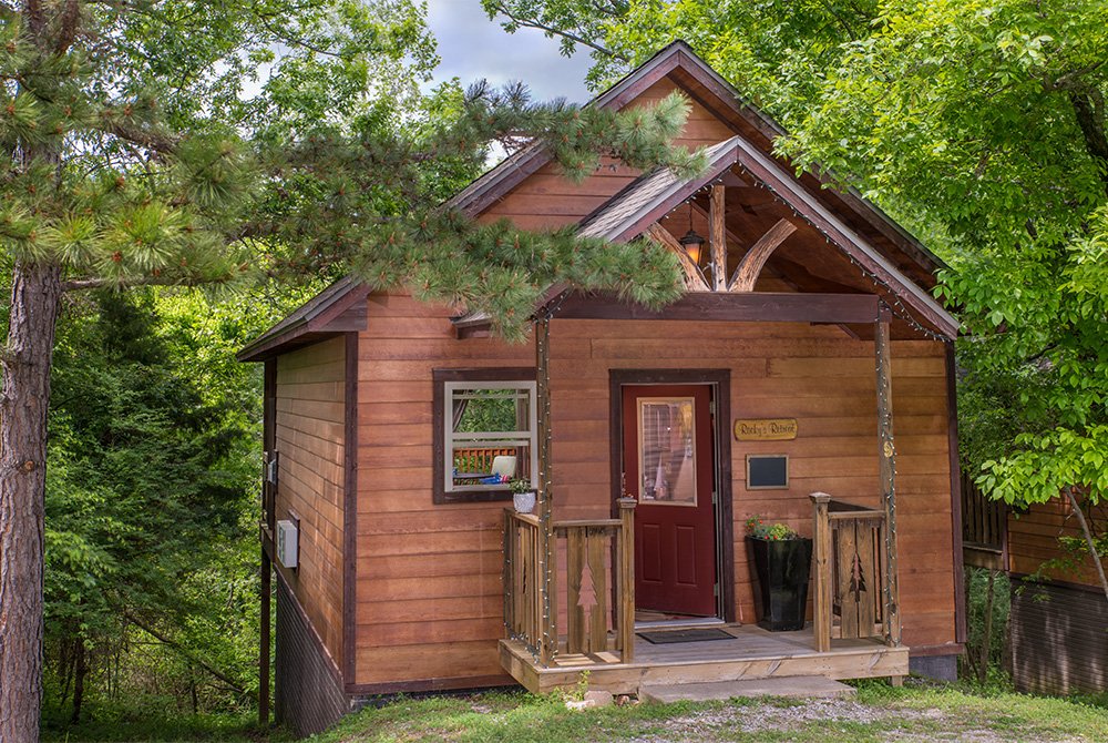 Exterior view of a small, rustic wooden cabin nestled among lush green trees, featuring a gabled roof and a small front porch with a dark red door.