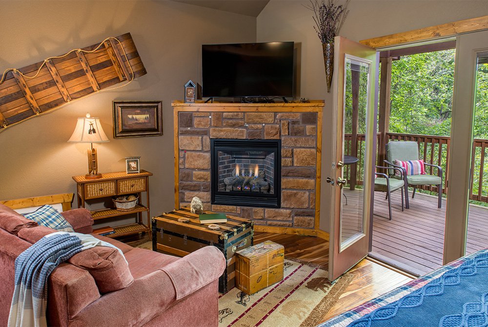 A cozy living area featuring a pink sofa with a blue throw blanket, a stone fireplace with a television mounted above it, and glass doors that open onto a wooden deck overlooking a forest.