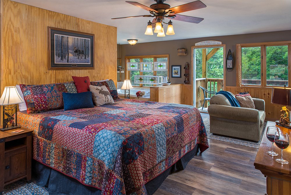 A spacious rustic bedroom featuring a king-sized bed with a colorful patchwork quilt, wood-paneled walls, a ceiling fan, and a view of a forested balcony through a glass door.