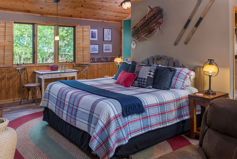 A cozy rustic bedroom featuring a king-sized bed with a blue and red plaid comforter, wood-paneled walls, and nautical-themed decor including wooden oars and a sled mounted above the headboard.