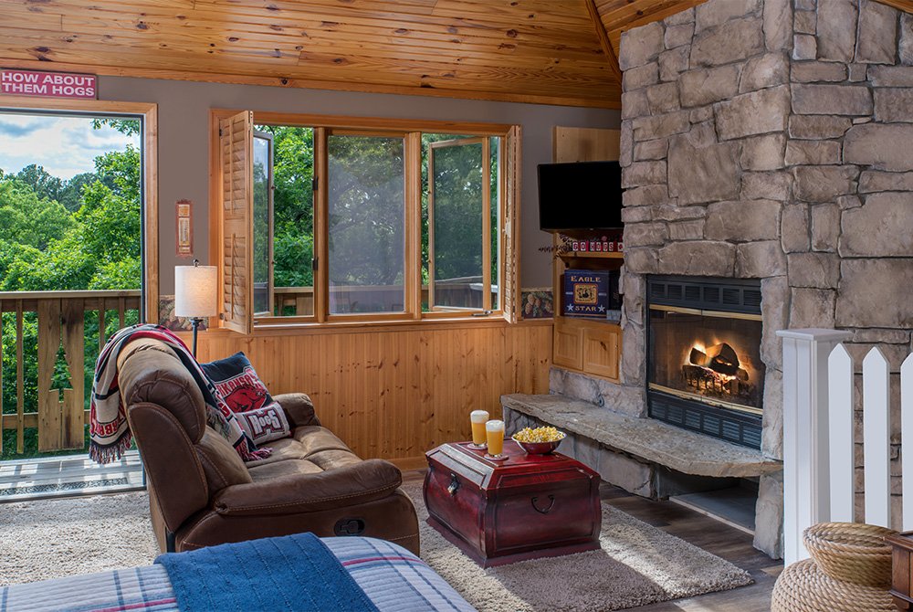 A cozy cabin living area featuring a brown leather recliner, a large stone fireplace with a glowing fire, and a wooden coffee table, with glass doors opening onto a balcony overlooking a lush green forest.