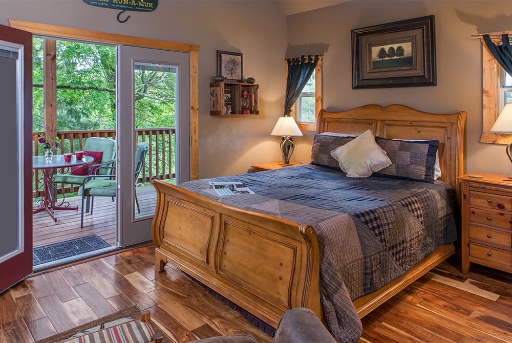 A cozy bedroom featuring a wooden queen sleigh bed and hardwood floors, with glass doors opening onto a private deck overlooking a lush green forest.