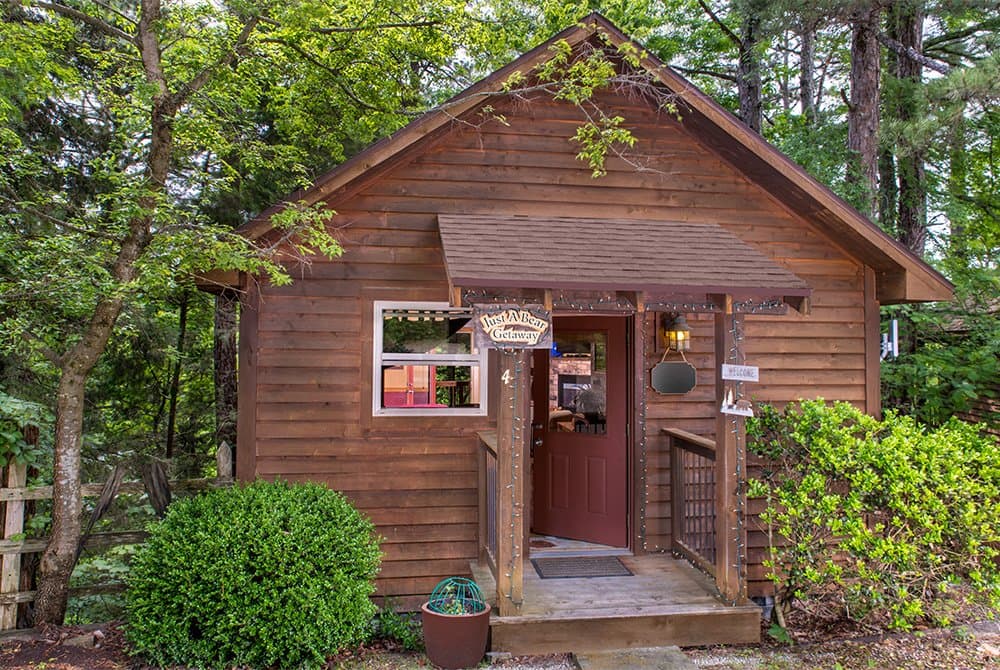 A rustic wooden cabin with a gabled roof and a small covered porch, surrounded by lush green trees and professional landscaping.