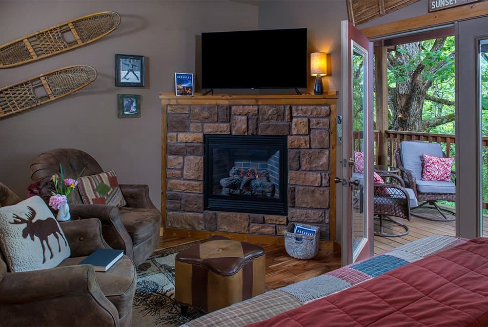 Cozy living area in a rustic cabin featuring a stone fireplace with a mounted flat-screen TV, comfortable leather armchairs with moose-themed pillows, and an open door leading to a scenic wooded balcony.