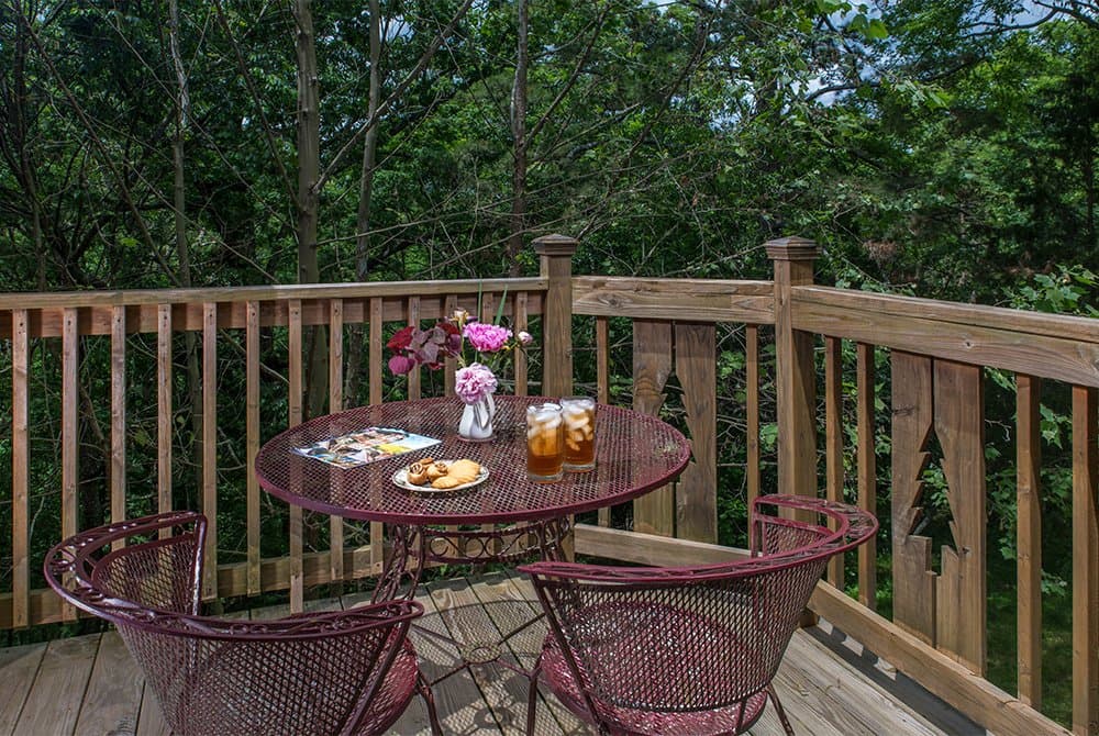 An outdoor wooden balcony featuring a round red metal table with two matching chairs, set against a backdrop of dense green trees.