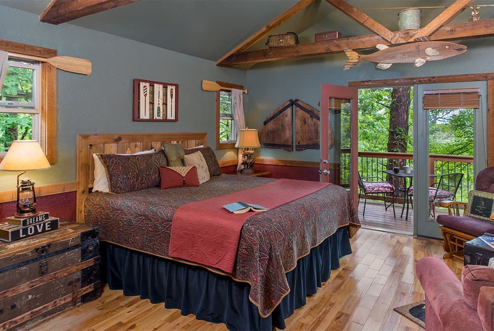 Bedroom interior with a king-size bed, featuring rustic fishing-themed decor like mounted oars and a large fish sculpture, with glass doors leading to a wooded balcony.