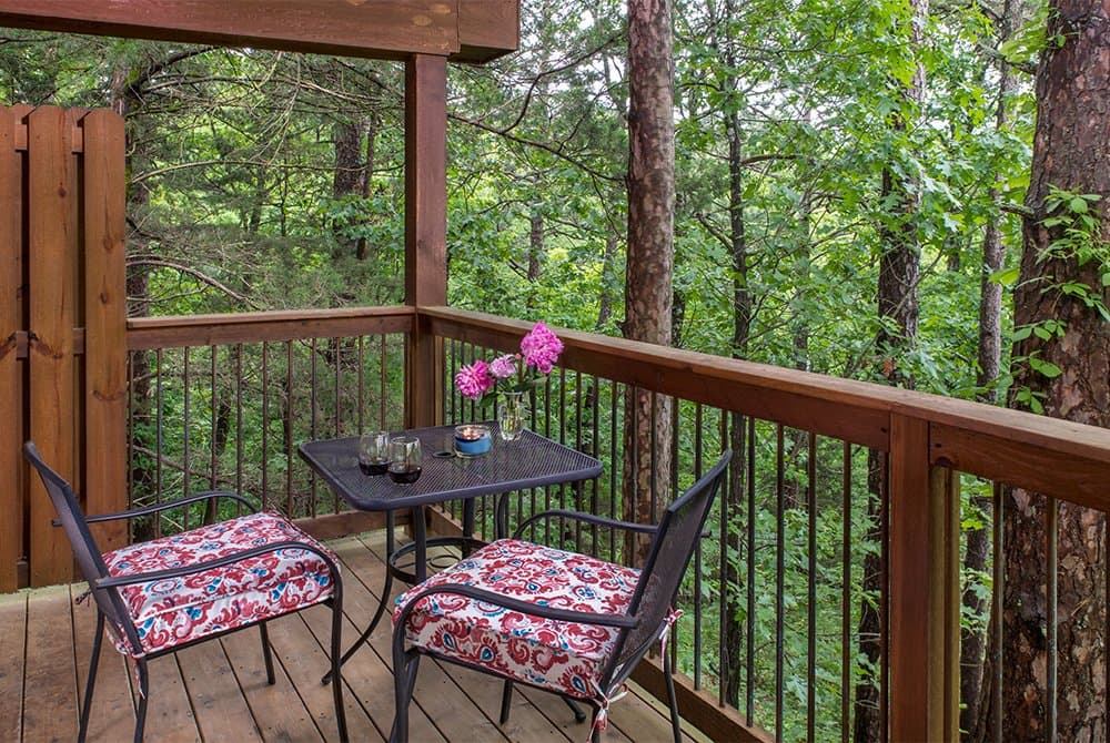 A wooden balcony deck with a small bistro table and two cushioned chairs, overlooking a lush green forest.