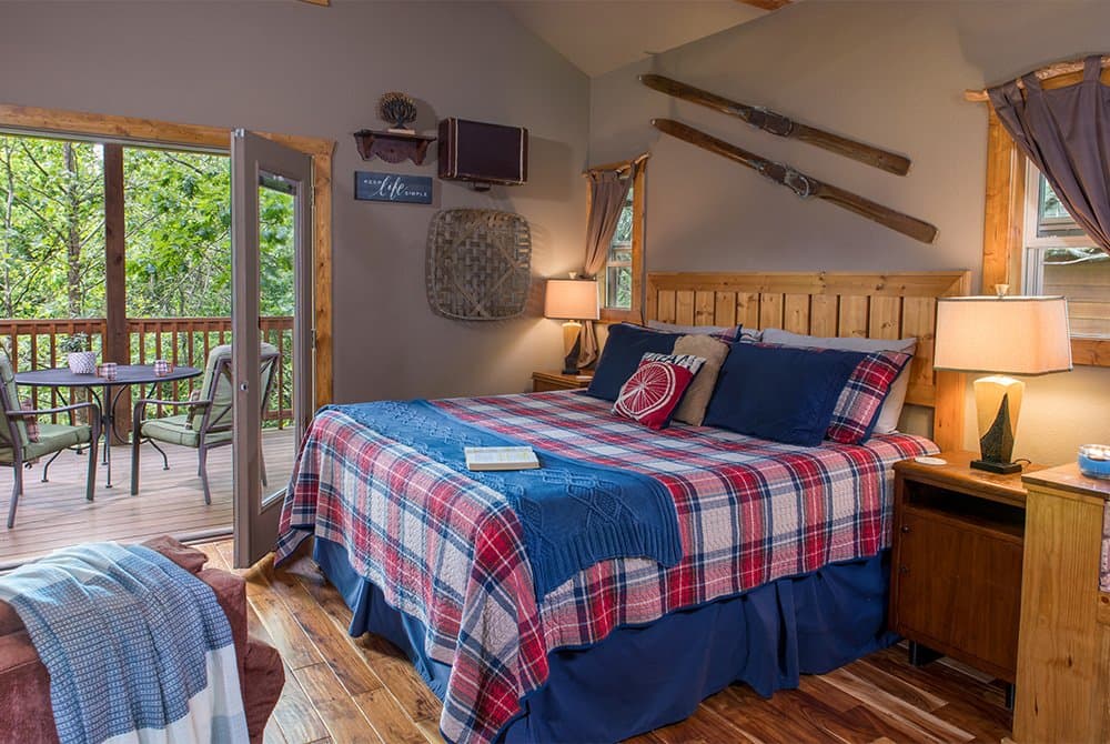 A rustic bedroom with a king-sized bed featuring a red and blue plaid comforter, with oars mounted on the wall and glass doors opening onto a wooded balcony.