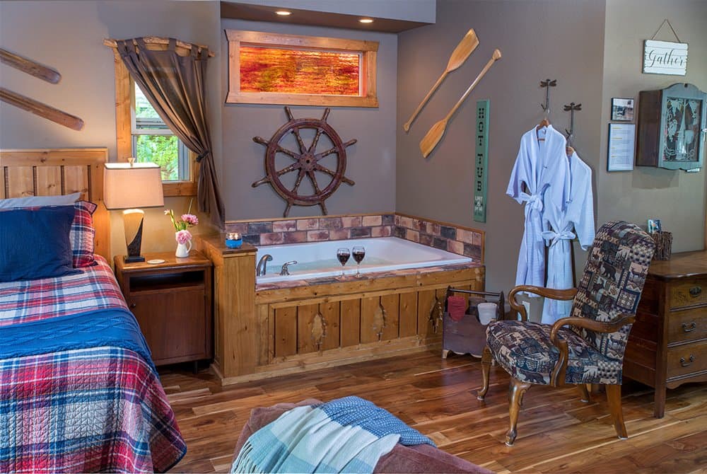 A cozy bedroom area featuring a king-sized bed with a red and blue plaid comforter and a large white Jacuzzi decorated with a ship's wheel and wooden oars on the wall.