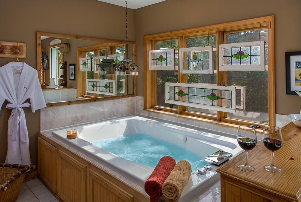 A double Jacuzzi filled with water and topped with wine glasses on the wooden edge, set against a window decorated with several small stained-glass panels.