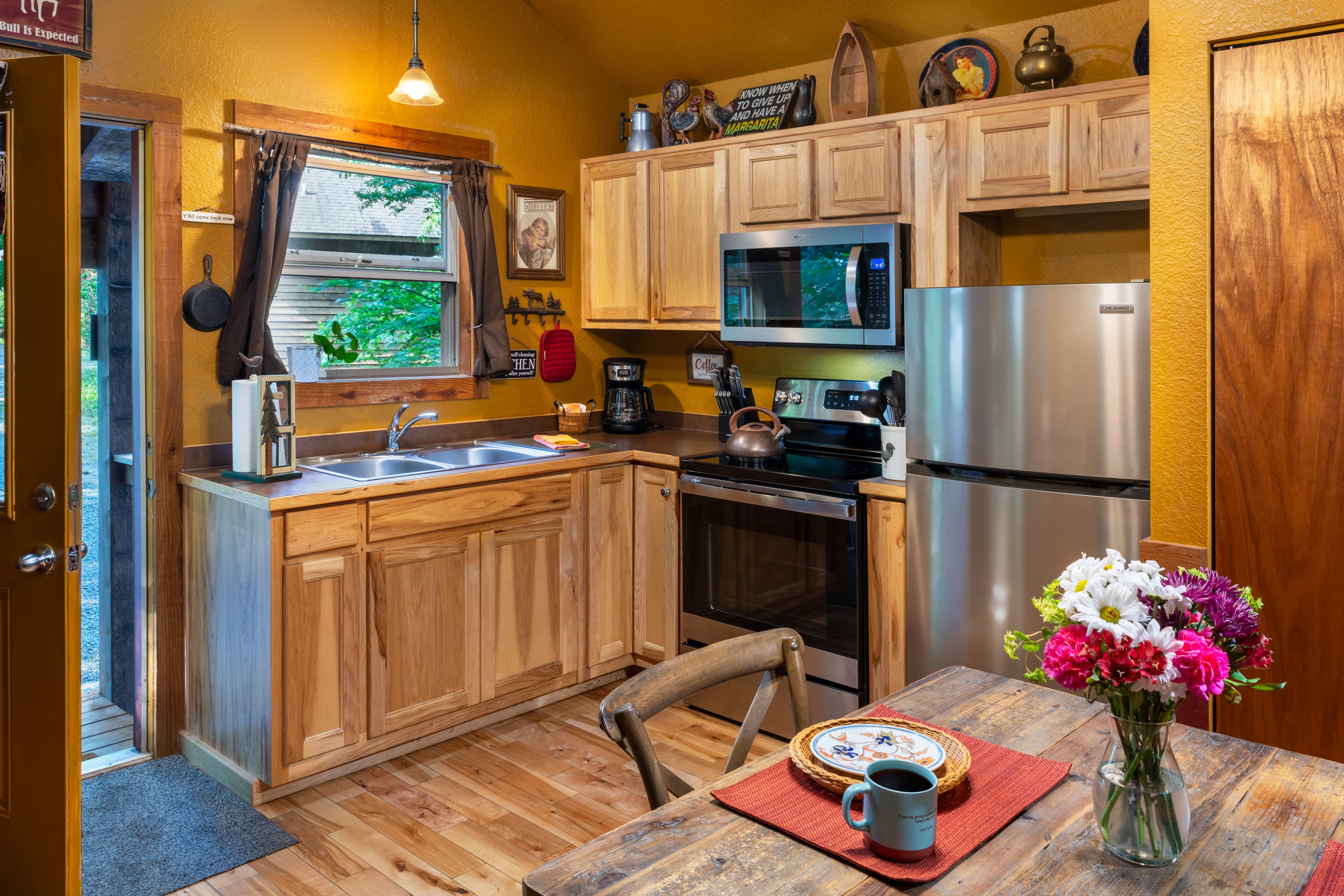 A fully equipped kitchen in a rustic cabin featuring stainless steel appliances, light wood cabinetry, a dining table with fresh flowers, and a window overlooking lush green trees.