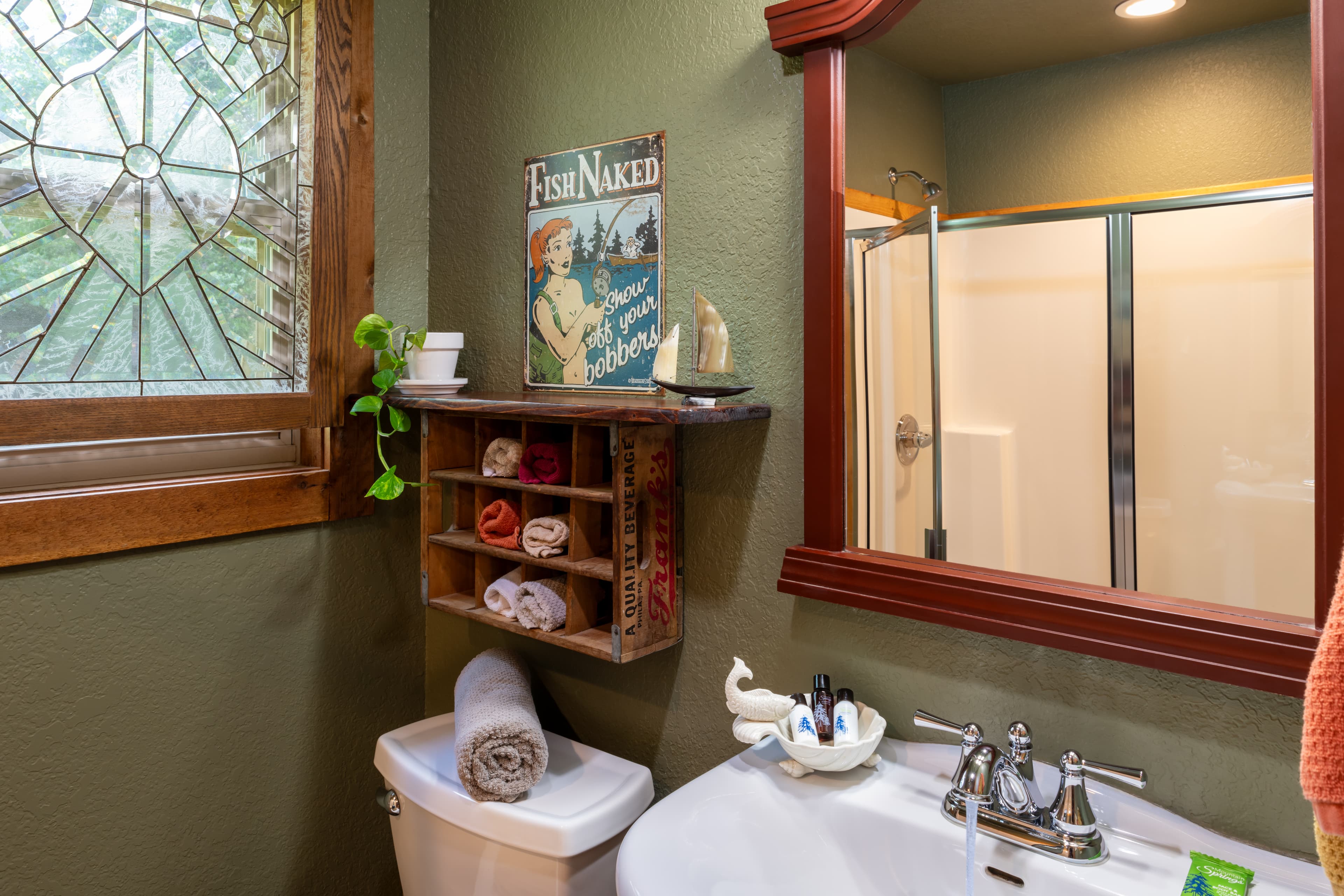 A rustic bathroom featuring olive green walls, a wooden shelf with rolled towels, a stained-glass window, and a white sink reflected in a wood-framed mirror.