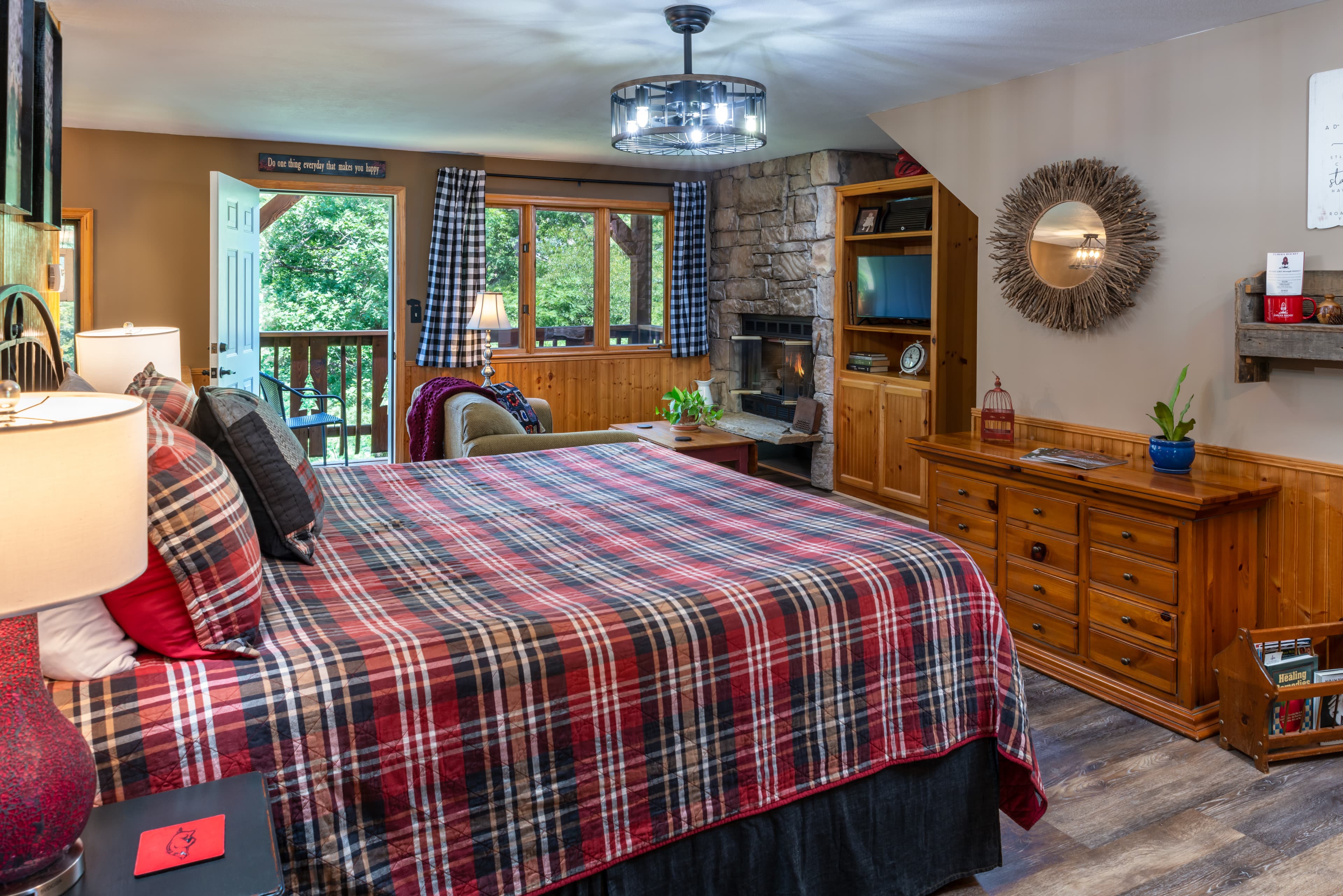 A spacious rustic bedroom with a king-sized bed featuring a red and grey plaid comforter, a stone fireplace with a built-in TV cabinet, and a glass door leading to a wooded balcony.