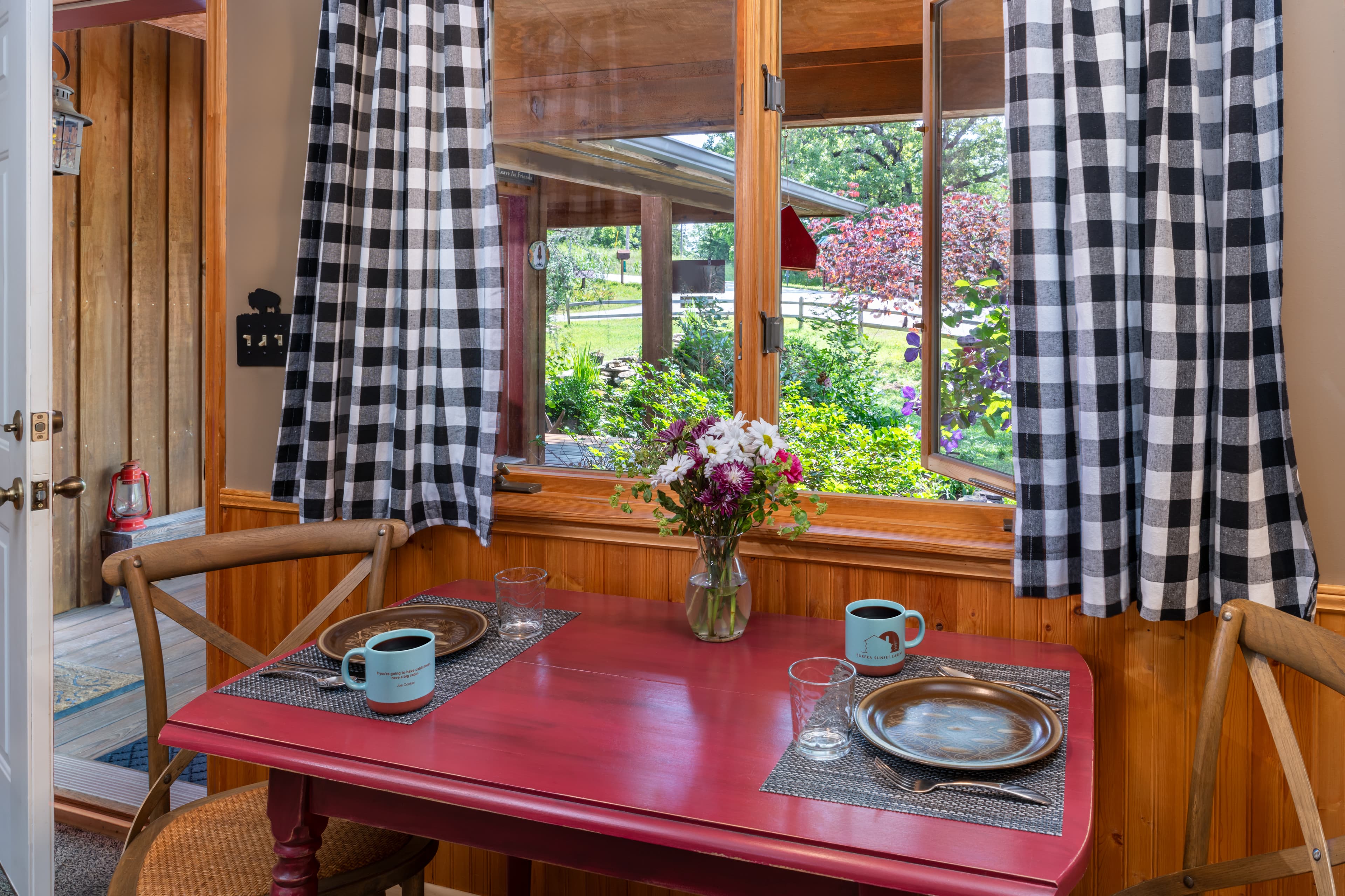 A simple dining area in a rustic cabin featuring a red wooden table set for two with blue mugs, cross-back chairs, and a window with black-and-white checkered curtains overlooking a green yard.