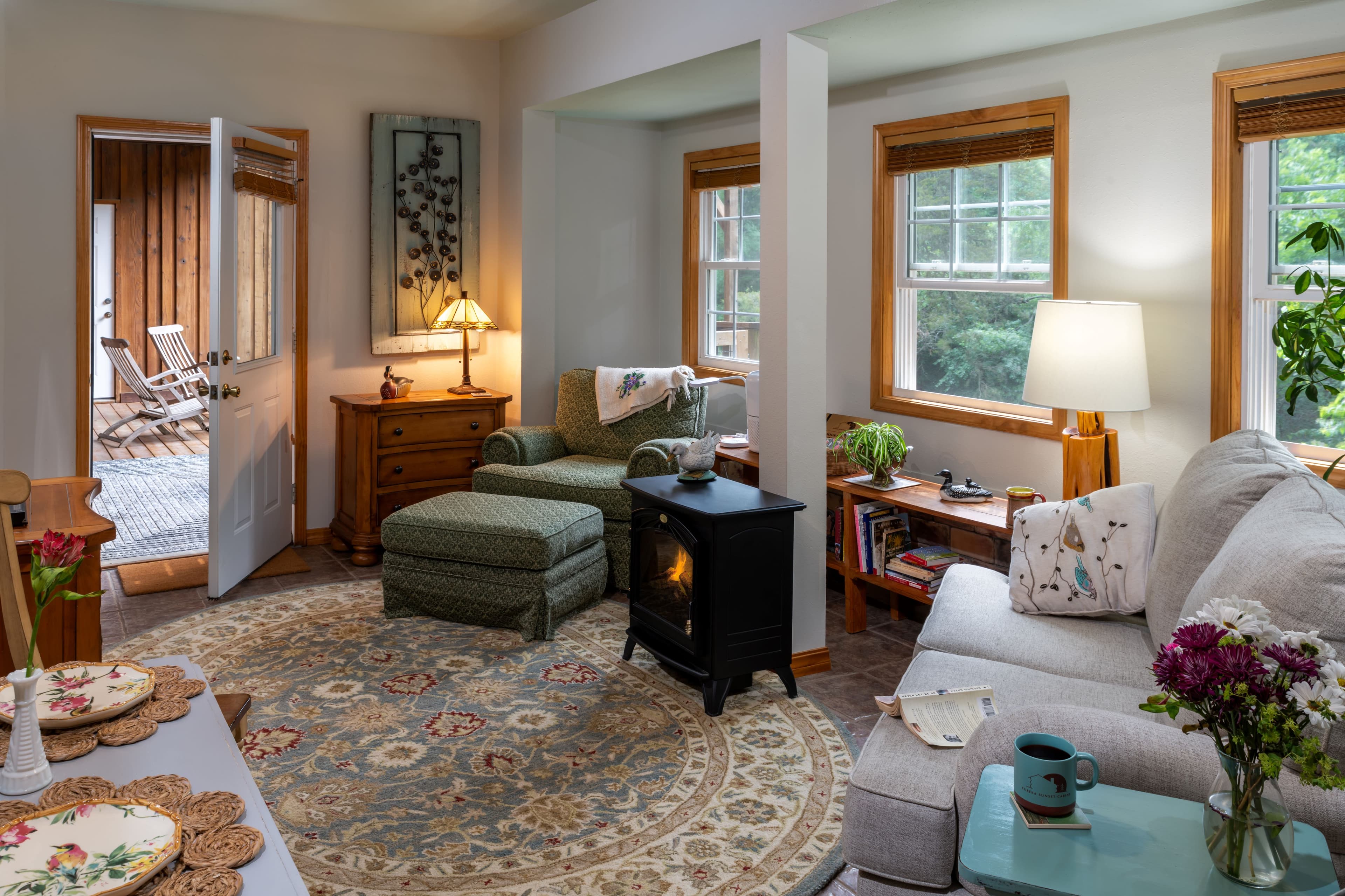 A bright and inviting living room in a rustic cabin featuring a white sofa, a green armchair with a matching ottoman, a small black electric stove, and large windows that fill the space with natural light.