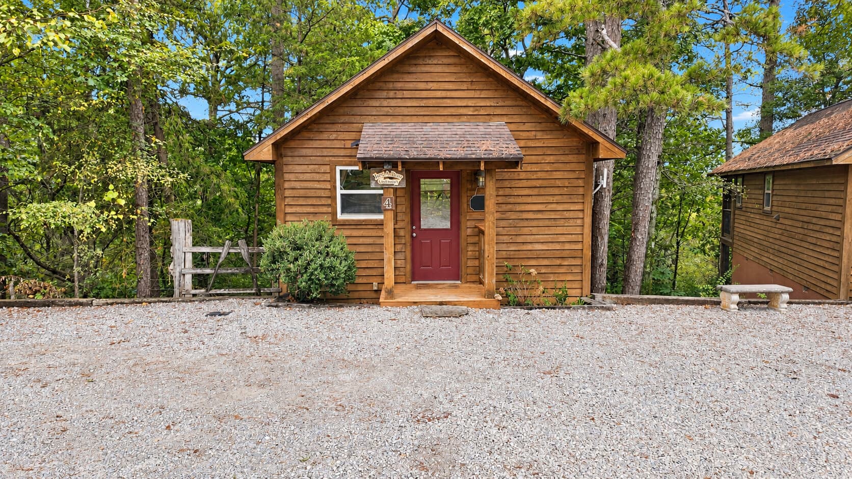 A cozy wooden cabin surrounded by trees with a gravel parking area in front.