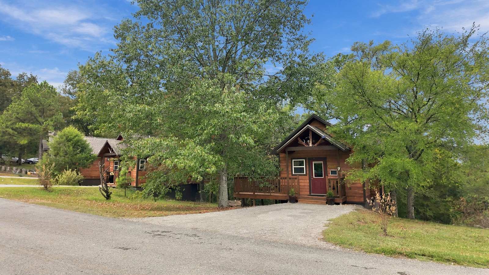 A wooden cabin surrounded by trees and greenery, featuring a gravel driveway.