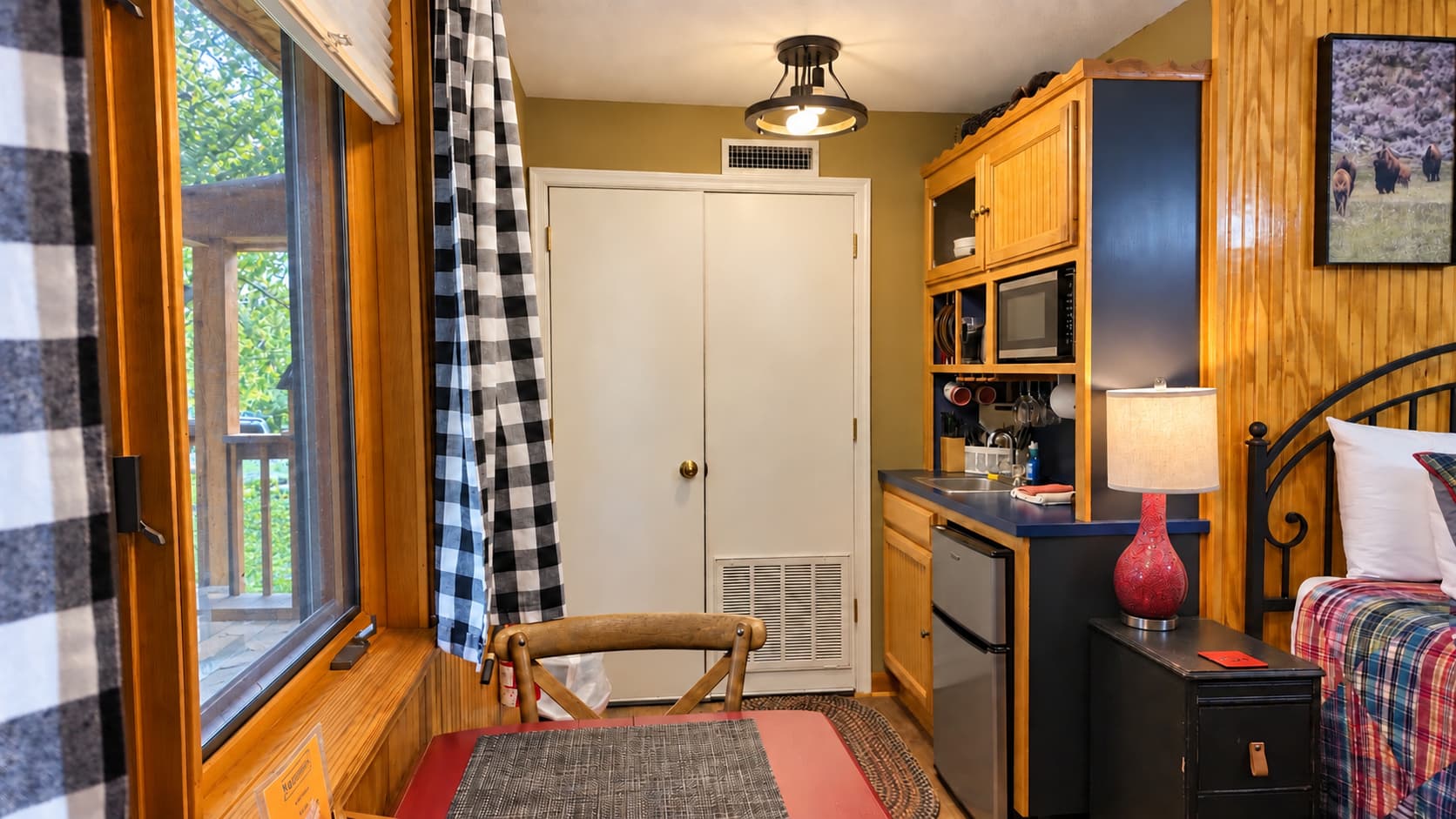 A cozy interior of a small living space featuring a kitchenette, dining area, and a view through a window with checkered curtains.