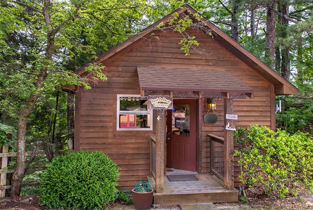 A rustic wooden cabin with a gabled roof and a small covered porch, surrounded by lush green trees and professional landscaping.
