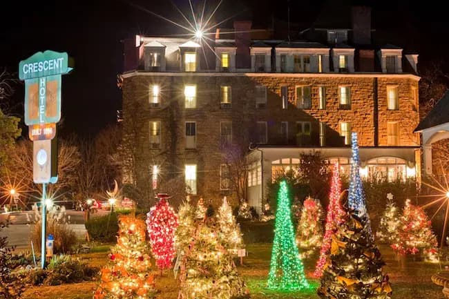A historic hotel illuminated by festive lights and surrounded by decorated Christmas trees at night.