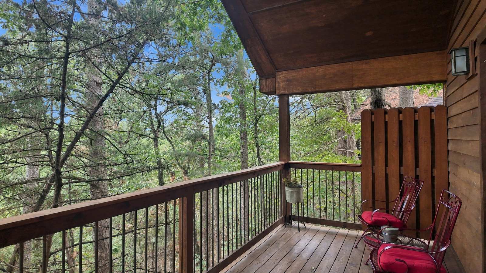 A wooden balcony with two red chairs overlooks a lush, green forest.