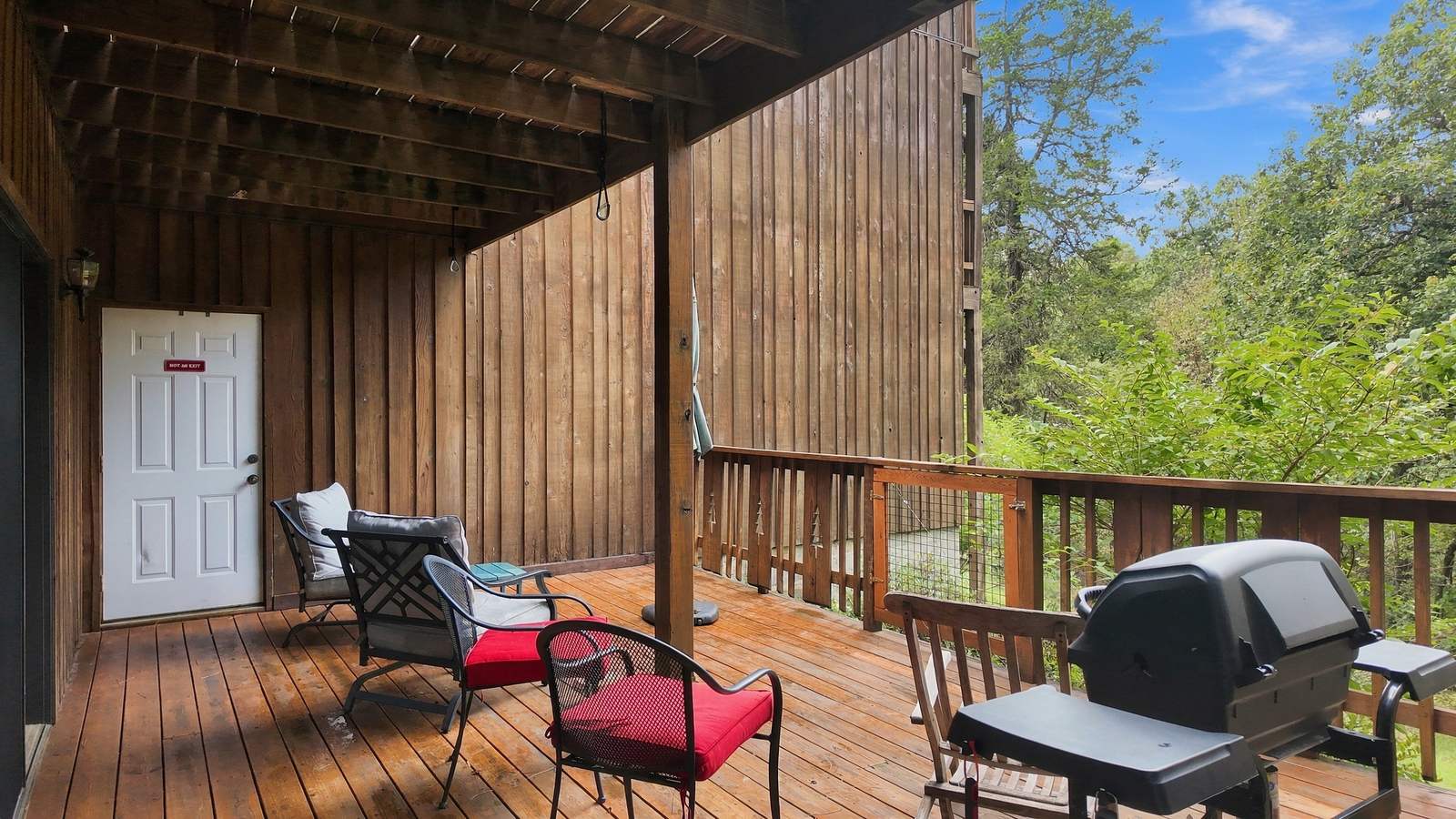 A wooden deck with chairs, a grill, and a white door surrounded by greenery.