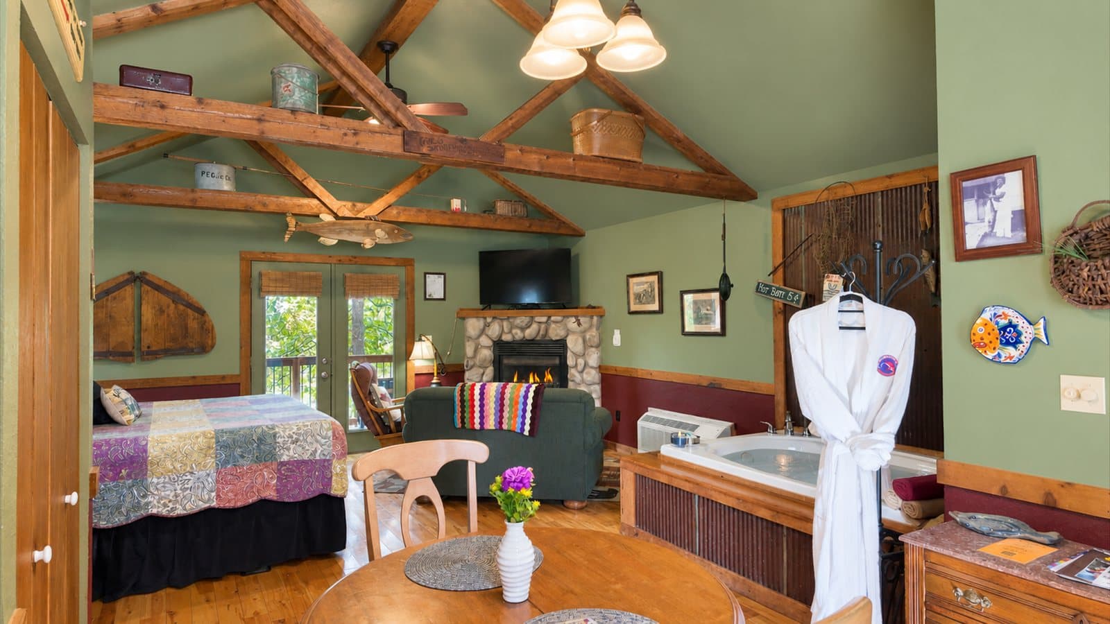 A cozy lodge interior featuring a bed, a seating area, a fireplace, and a hot tub, with rustic wooden beams and green walls.