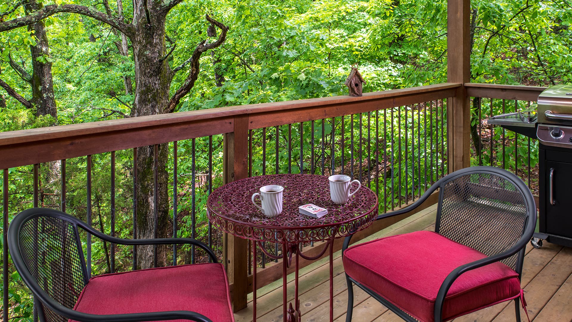 A cozy balcony features a small round table with two mugs and red cushioned chairs, surrounded by lush greenery.