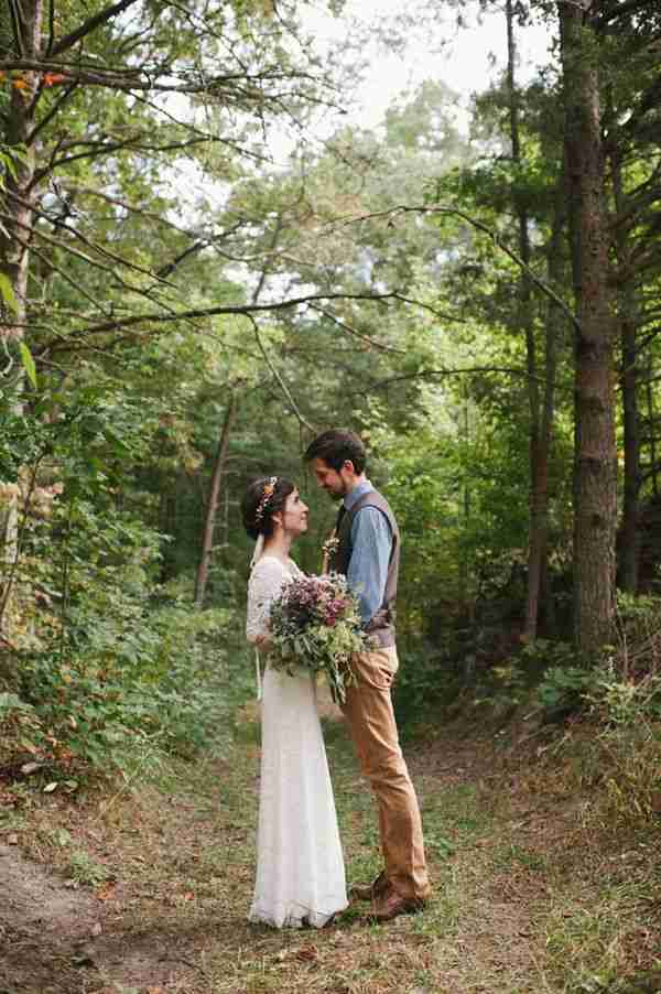 A bride and groom stand together in a wooded path, both gazing lovingly at each other.