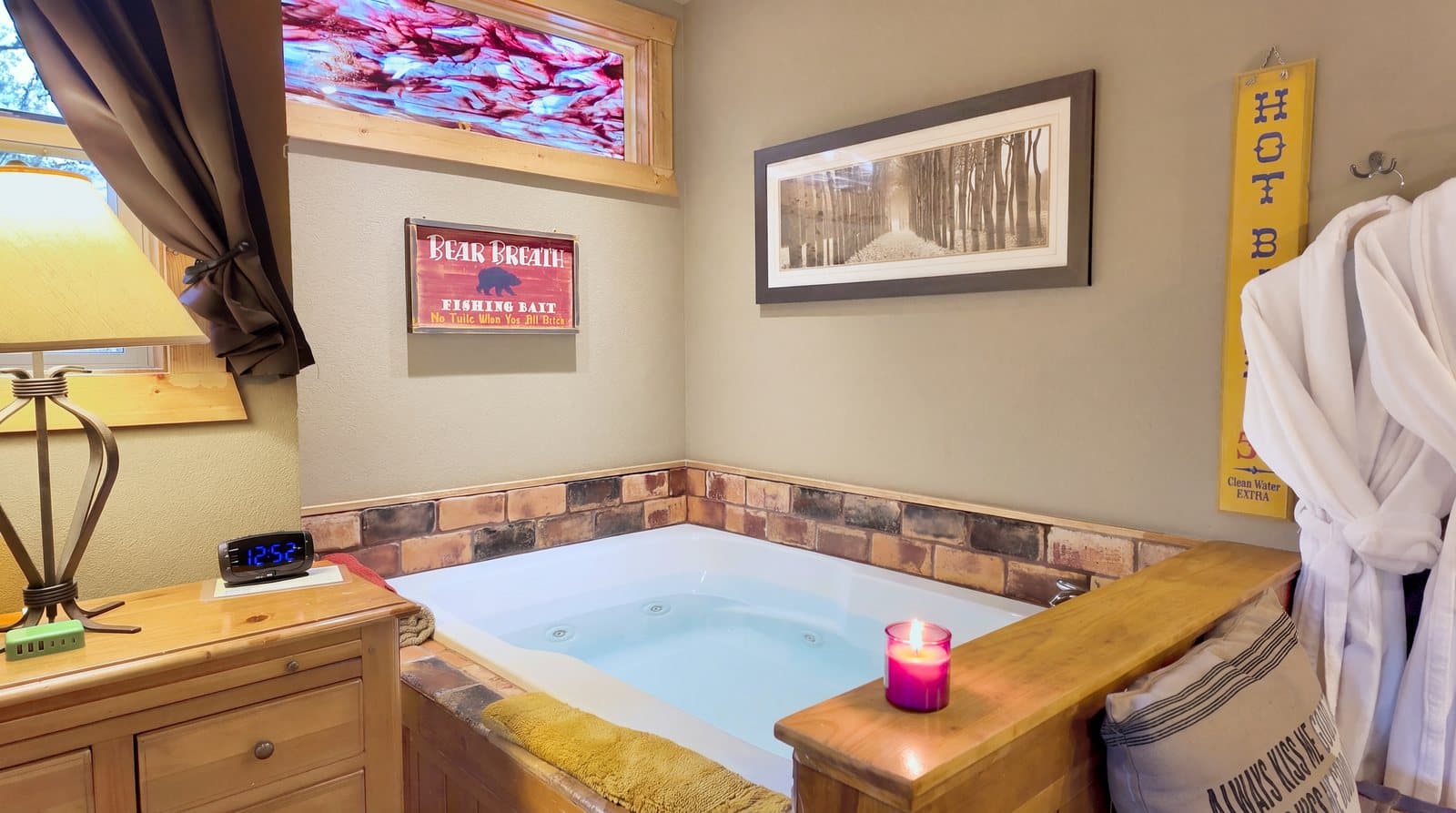 A cozy bathroom with a hot tub, lamp, and wooden furnishings, featuring a candle and decorative wall art.