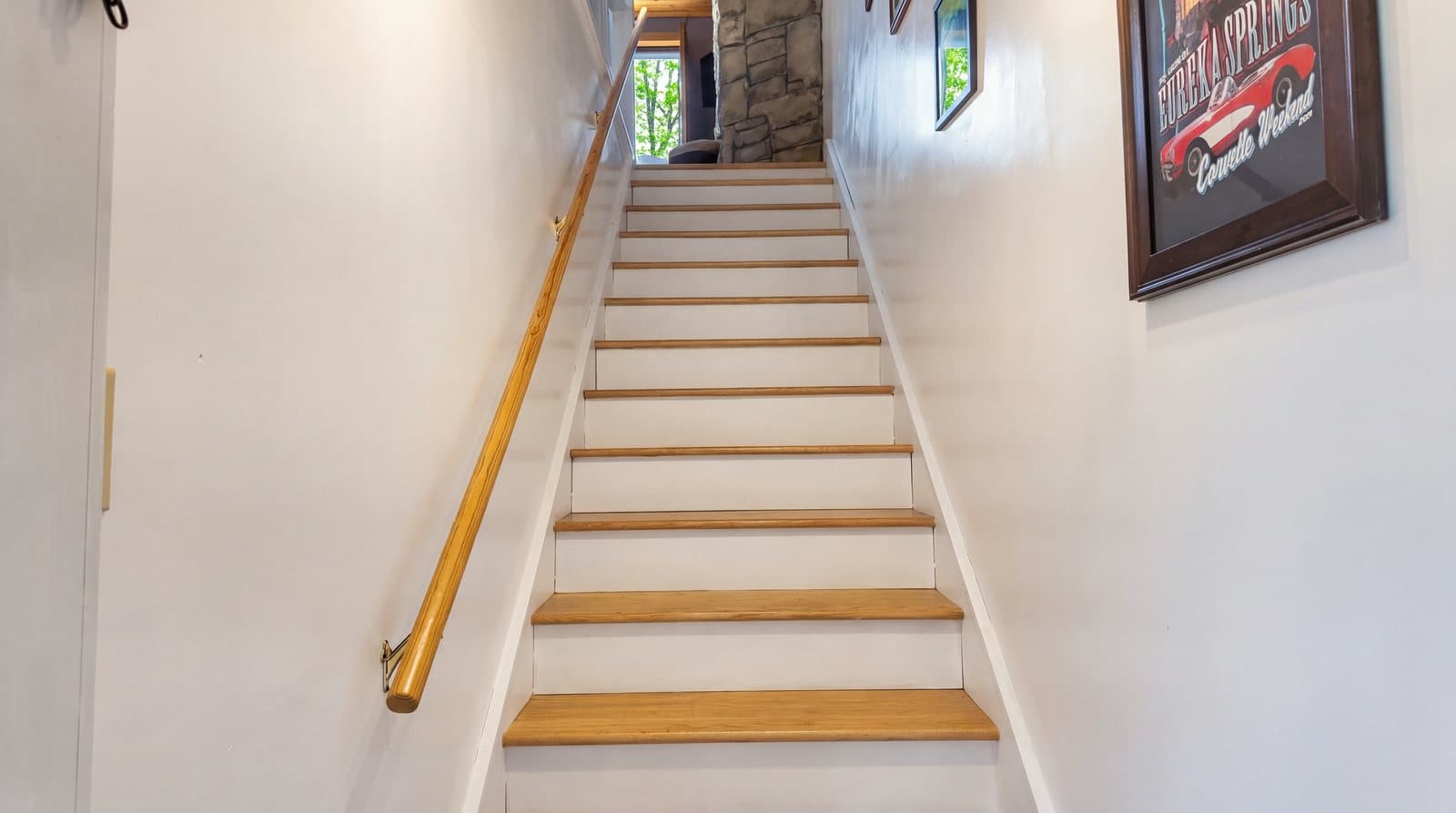 A set of white stairs with wooden treads leading upward, flanked by a wall and framed artwork.