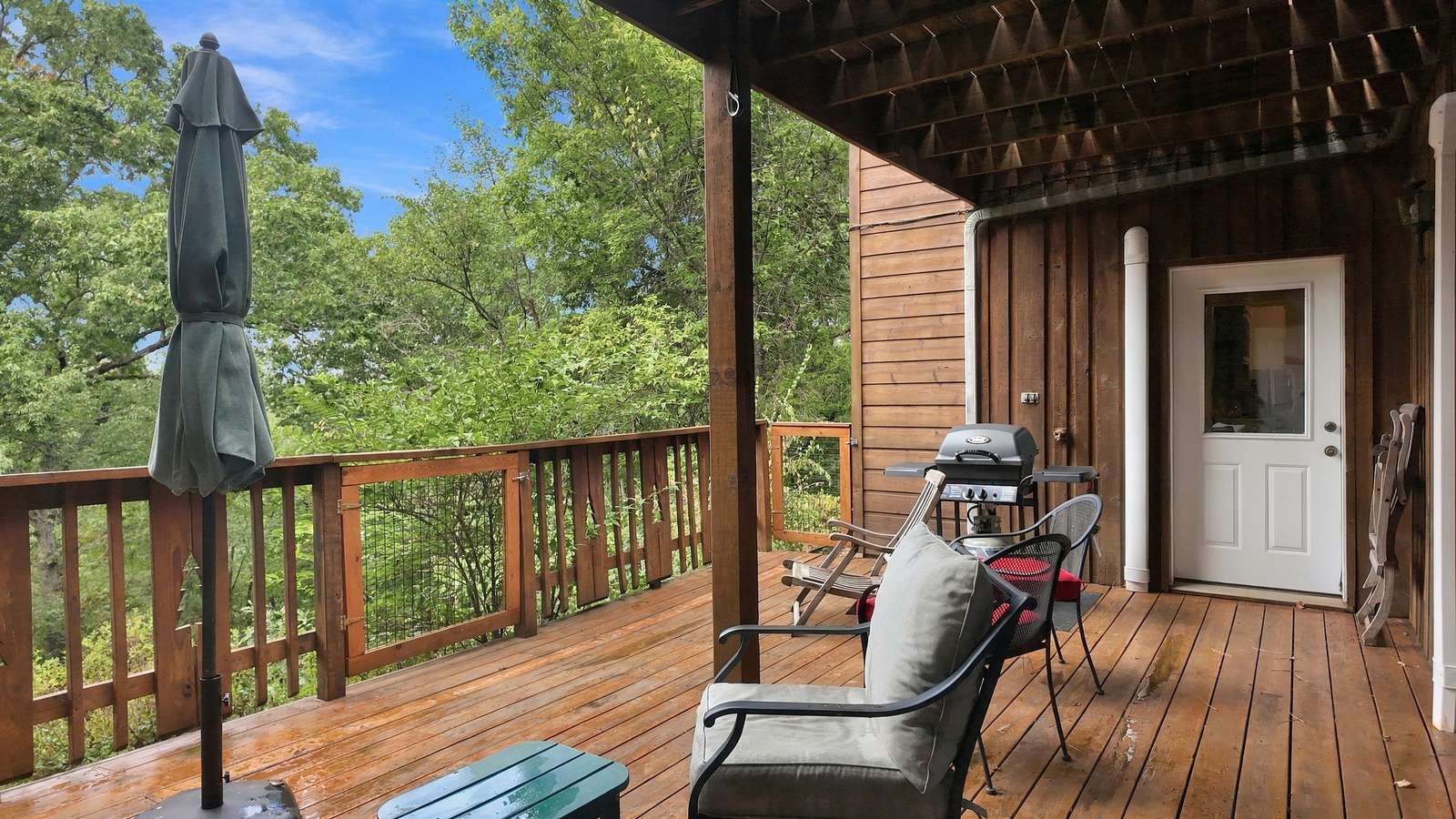 A wooden deck with chairs, a table, a grill, and a view of greenery.