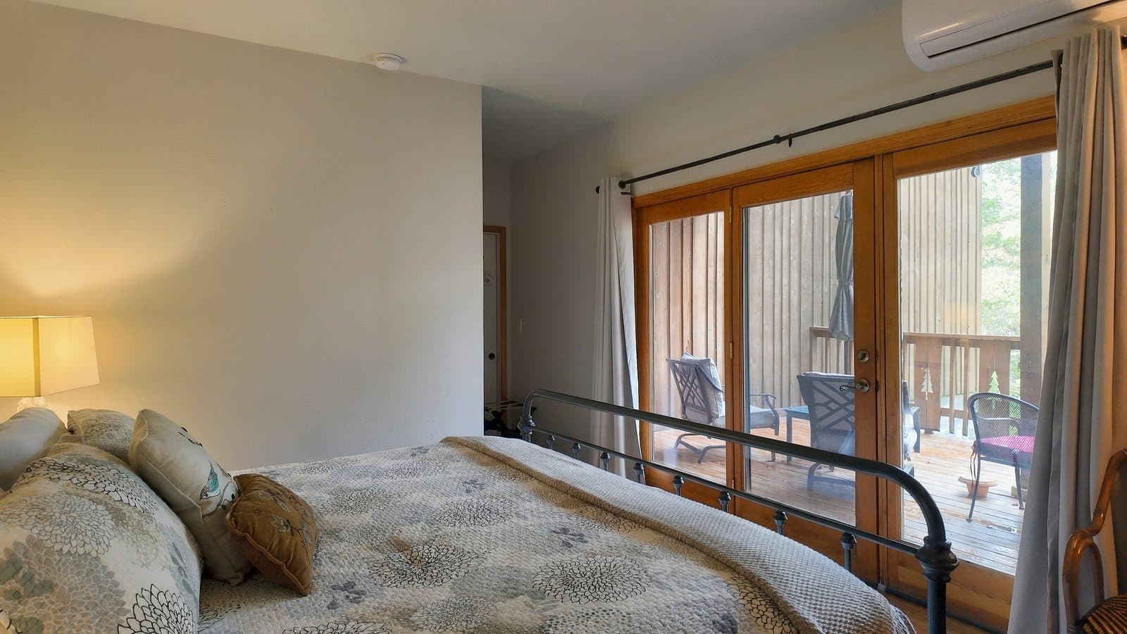 A cozy bedroom featuring a bed with decorative pillows, a bedside lamp, and large sliding doors opening to a wooden deck.