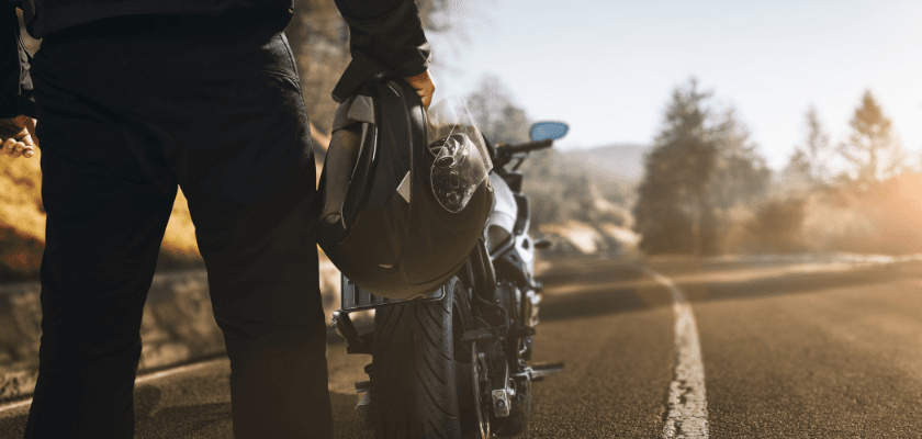 A motorcyclist holding a helmet stands next to their bike on an empty road surrounded by trees. A motorcyclist holding a helmet stands next to their bike on an empty road surrounded by trees.