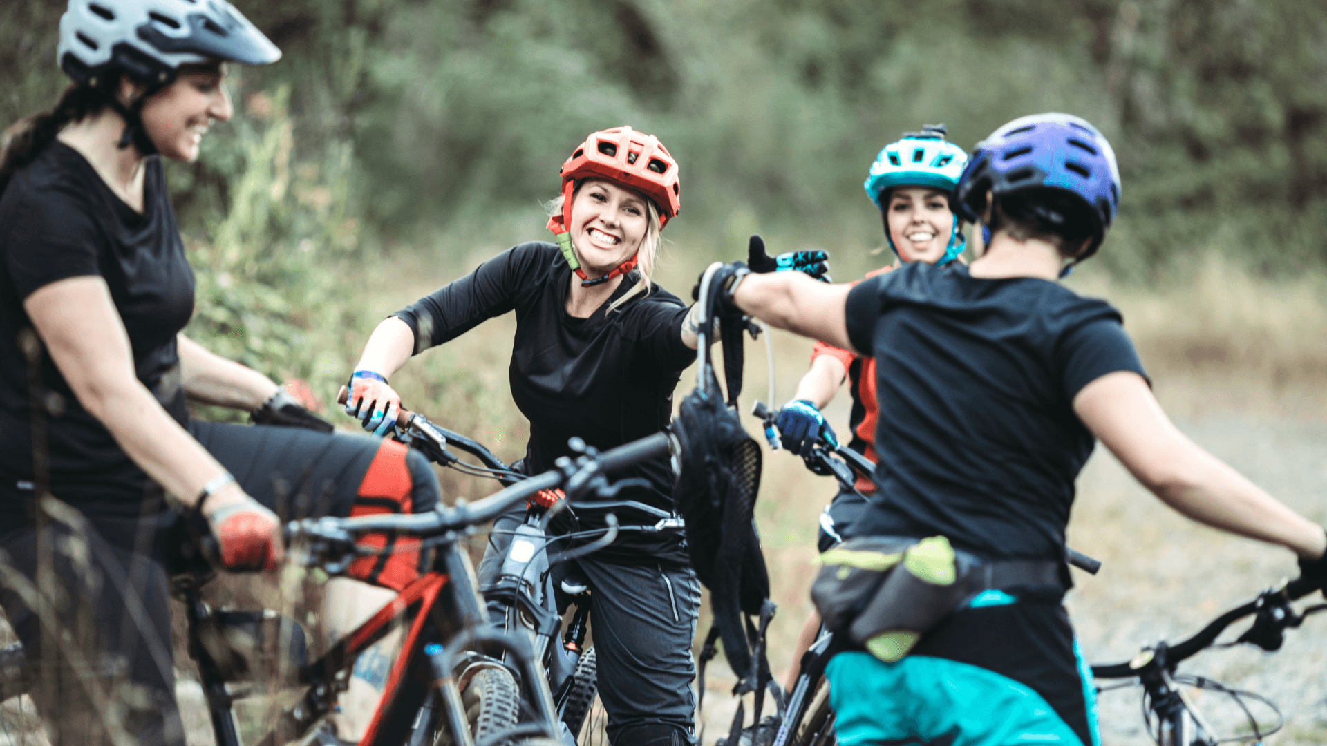 A group of four women wearing helmets and biking gear share a joyful moment outdoors.