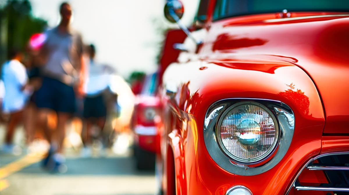 Close-up of a shiny red vintage car with blurred figures walking in the background. Close-up of a shiny red vintage car with blurred figures walking in the background.