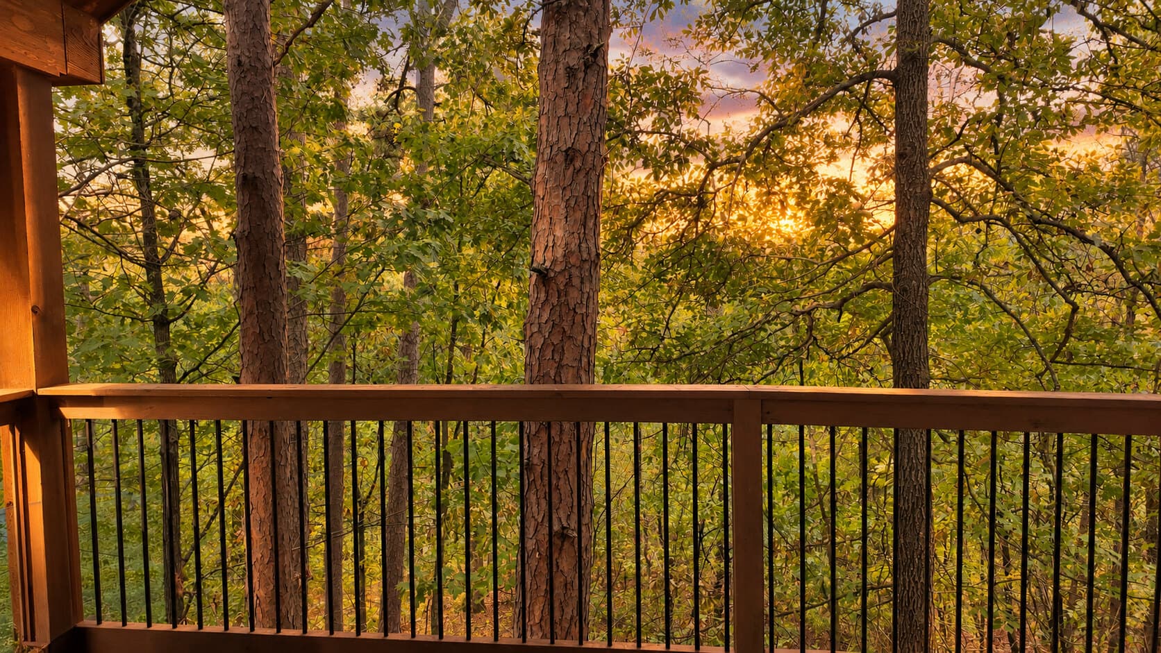 A wooden balcony railing overlooks a forested area at sunset.