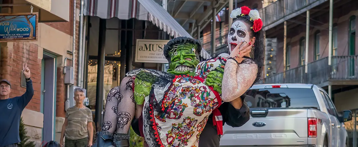 A man in a Frankenstein costume lifts a woman in Day of the Dead makeup and attire on a bustling street.