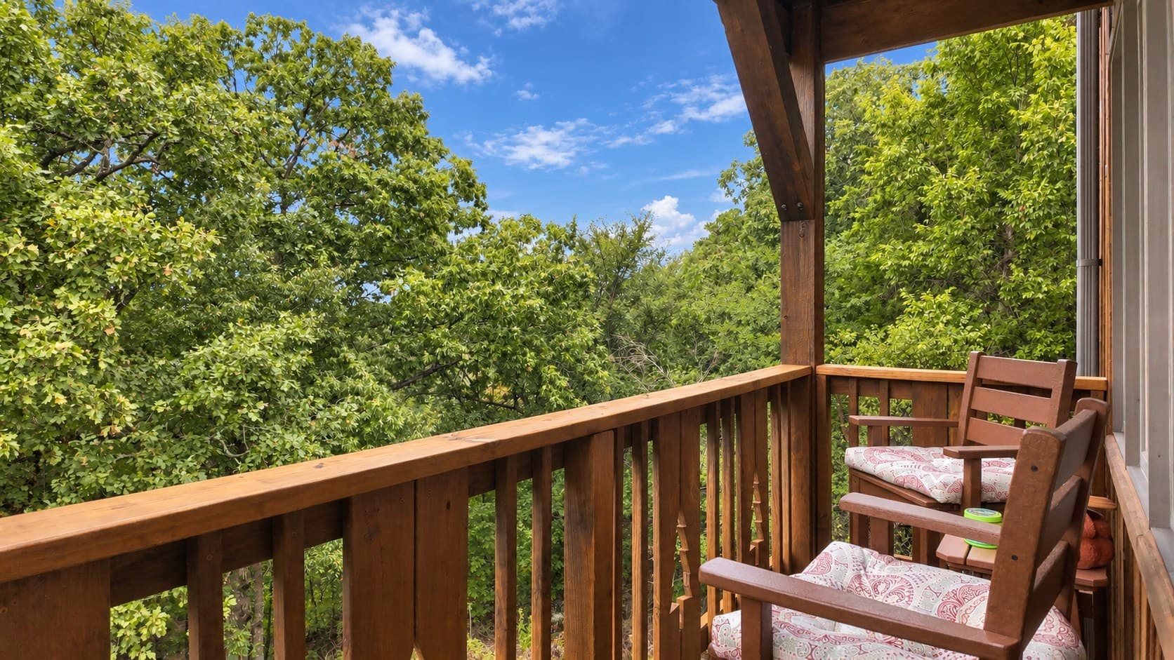 A wooden balcony with two chairs overlooks lush green trees under a blue sky.