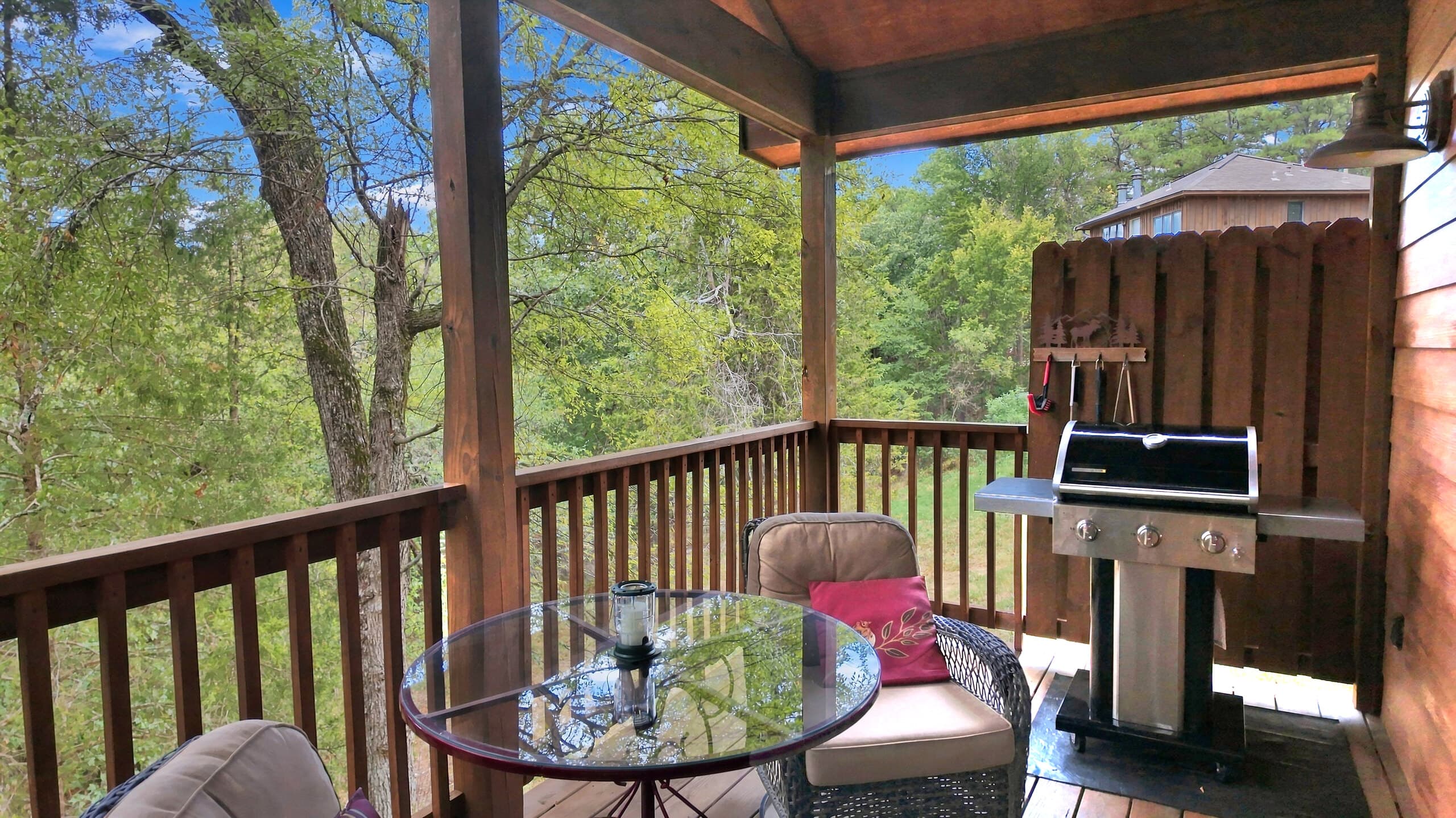 A covered balcony featuring a glass table, chairs, and a grill overlooking lush greenery.