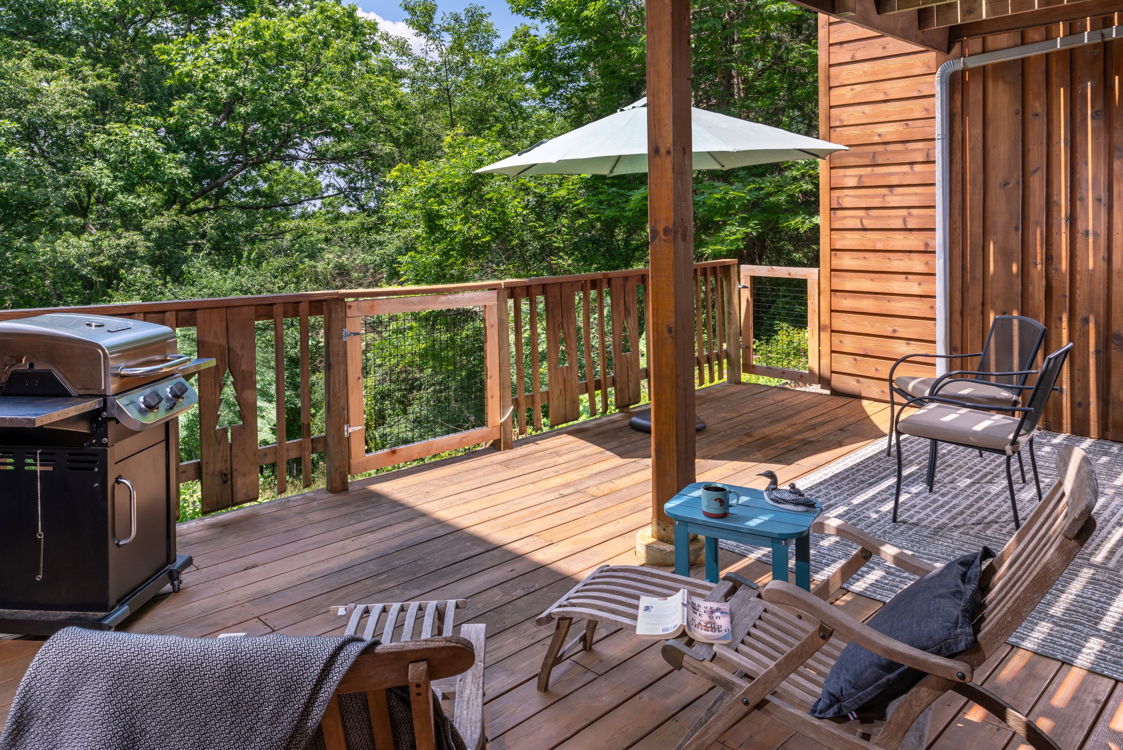 A spacious wooden deck with a gas grill, lounge chairs, a patio table under an umbrella, and a railing overlooking a dense green forest.