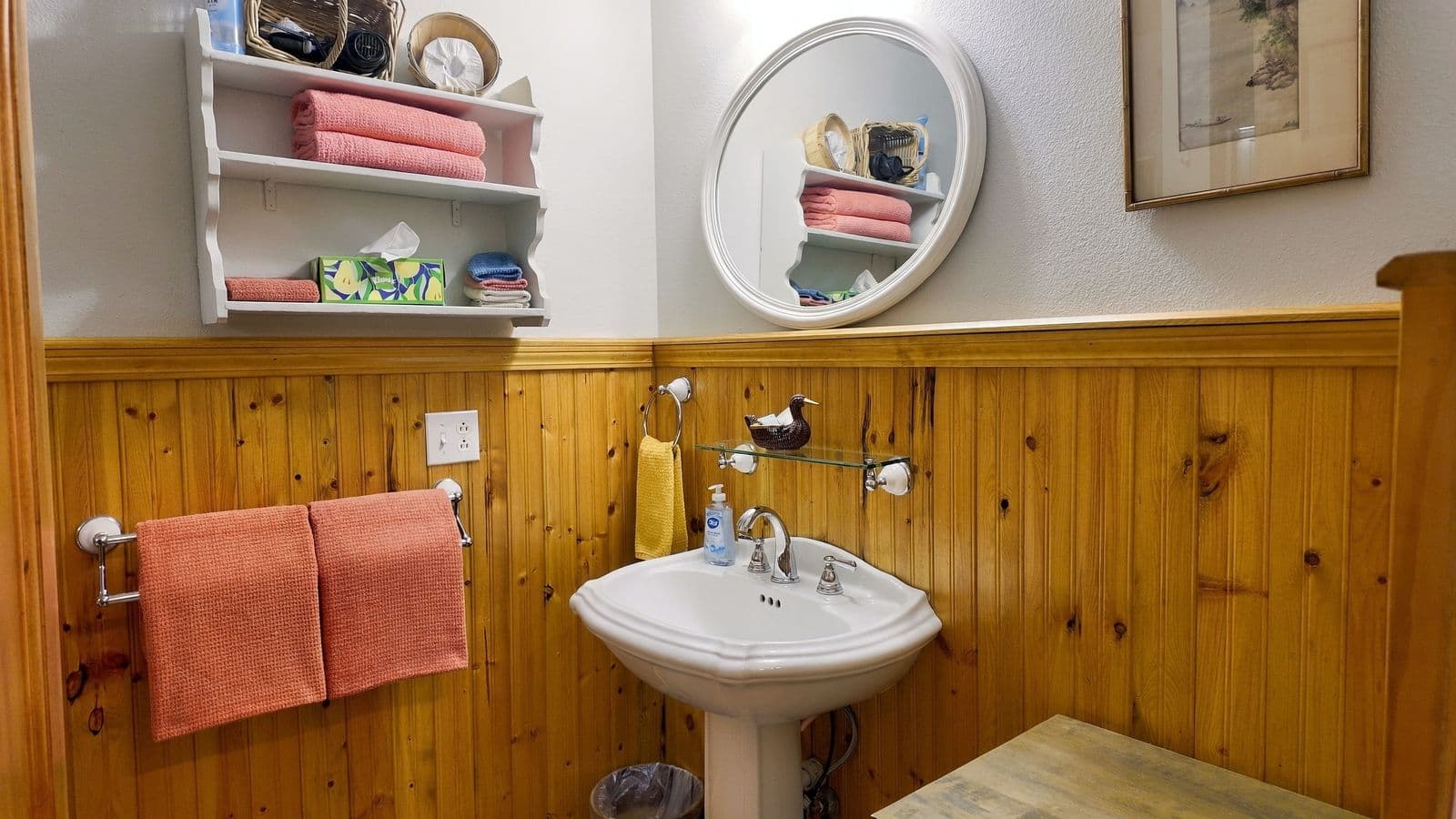A small bathroom featuring a white sink, a round mirror, wooden paneling, and neatly arranged towels on shelves.