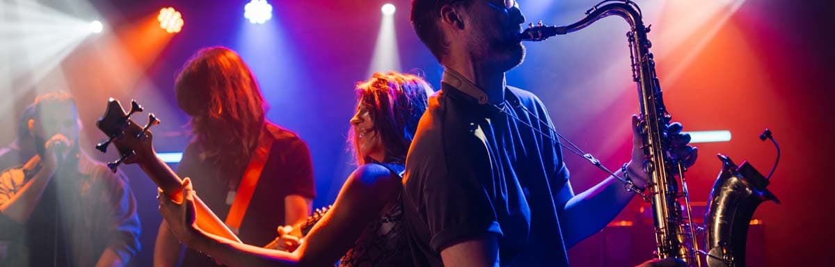 A musician plays saxophone on stage while other band members perform under colorful lights.