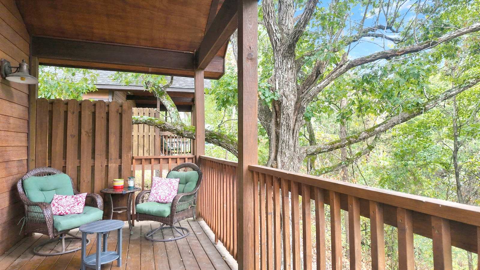 A cozy wooden balcony with two green chairs, a small table, and a view of trees.